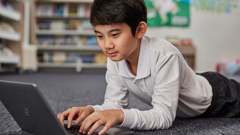 Image of young boy in polo shirt lying on floor and interacting with laptop device