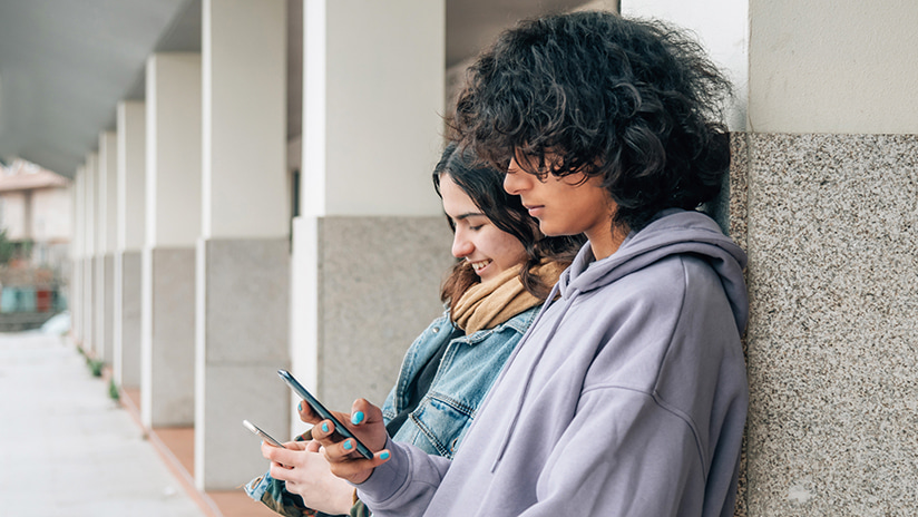 Photo of two young people using mobile phones and leaning on a wall at school