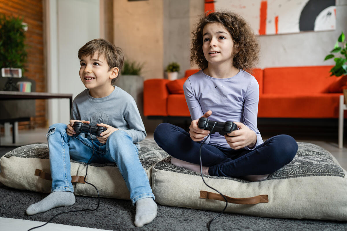 Two young children sitting on couch cushions at home while holding video game controllers and playing a video game at home