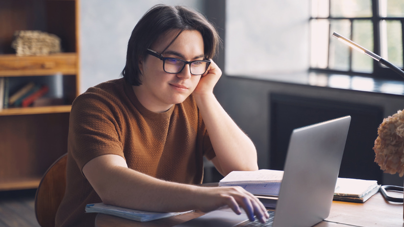 Photo of teenage boy with long hair and glasses working doing his homework on a laptop at the dining table at home